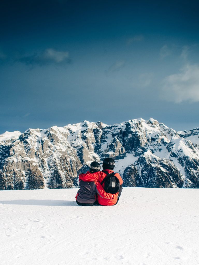 a couple sit hugging with their backs to the camera and facing a snow-covered mountain range at a ski resort in the US