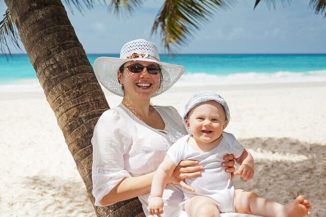 a baby sits smiling on a woman's lap as they both sit under a palm tree on a tropical beach