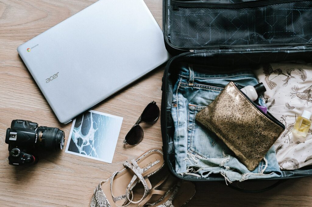 a packed suitcase lays open with a camera, sandals, sunglasses and a laptop outside of it