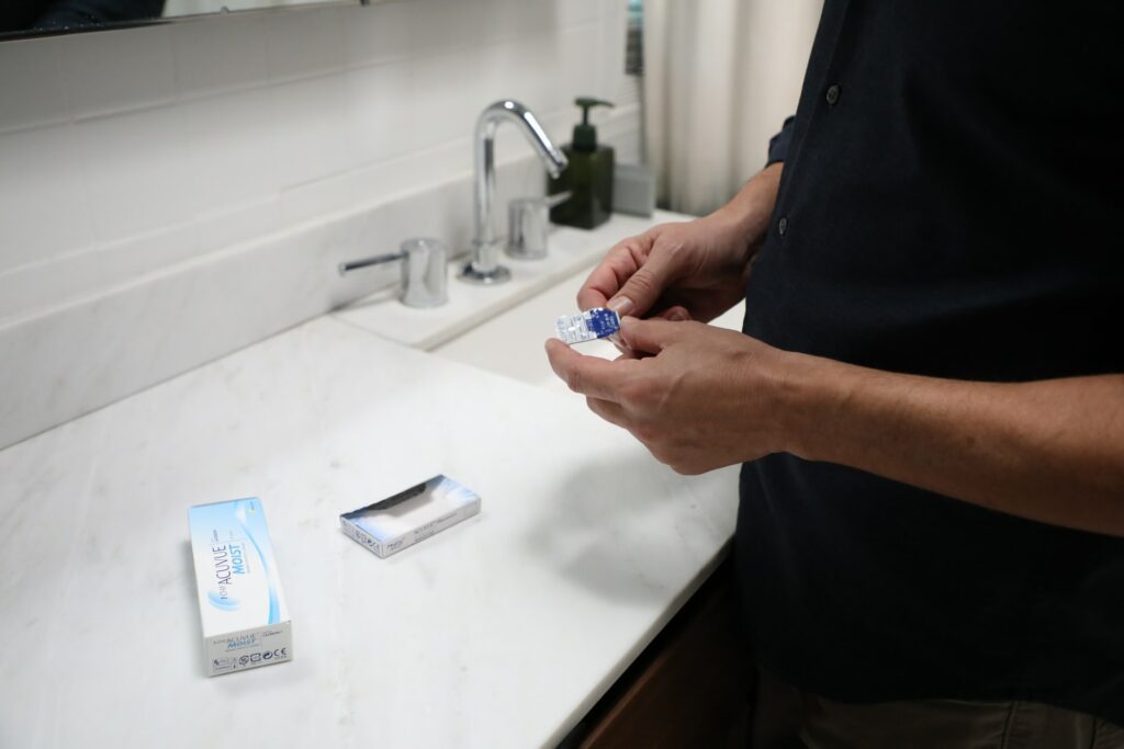 a mans stands at the bathroom counter looking at a package of contact lenses while the box sits on the white counter in front of him