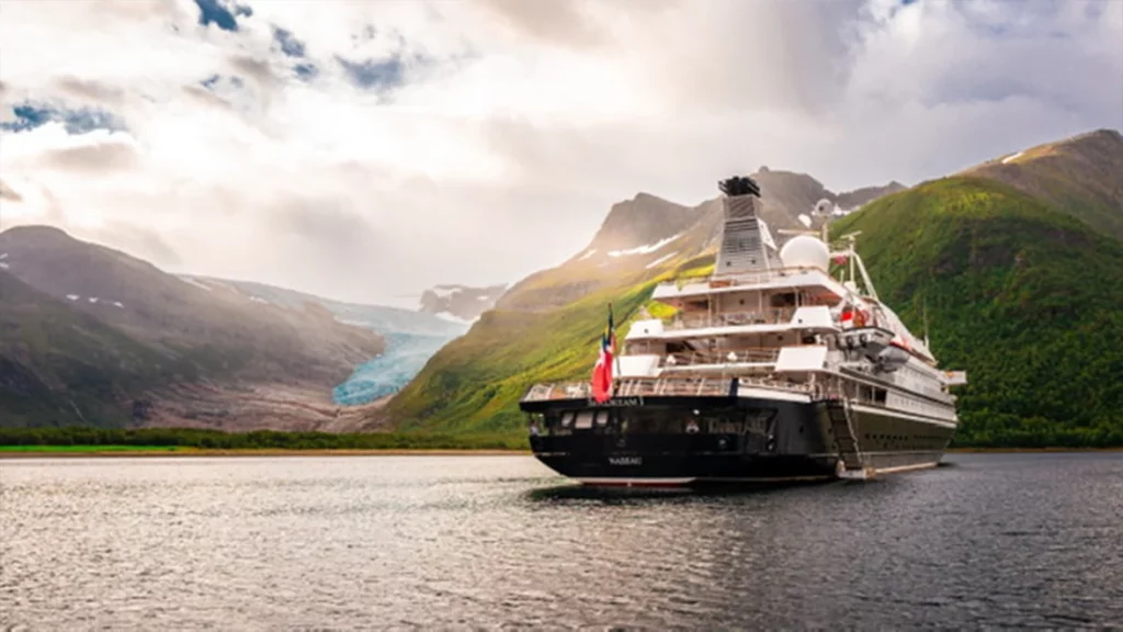 Luxury SeaDream yacht anchored in a serene Norwegian fjord with a glacier in the distance and lush green hills under a cloudy sky.