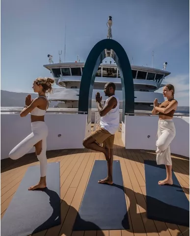 Passengers practicing yoga on the deck of a Swan Hellenic cruise ship, with clear skies and the ship's bridge in the background.