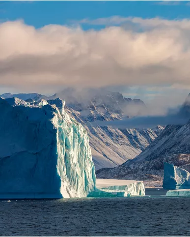 A towering iceberg rising majestically above the sea with a dramatic mountainous backdrop under a cloudy sky.